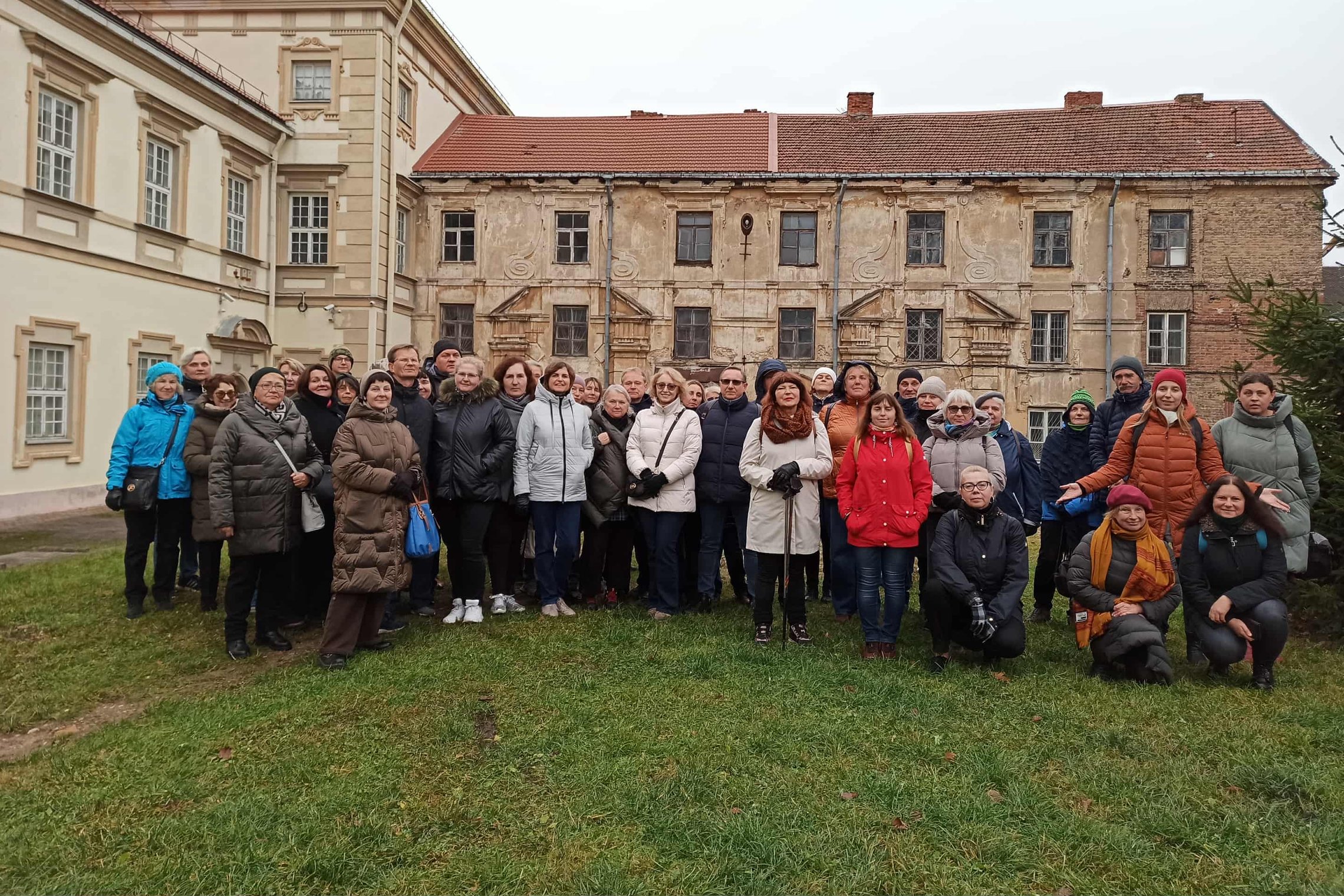 Didelė žmonių grupė stovi lauke priešais du istorinius pastatus. Kairėje matyti tvarkingas, šviesiai kreminės spalvos rūmas su raudonu stogu, o dešinėje – senesnis, nublukusios ir vietomis aptrupėjusios fasado sienos pastatas
