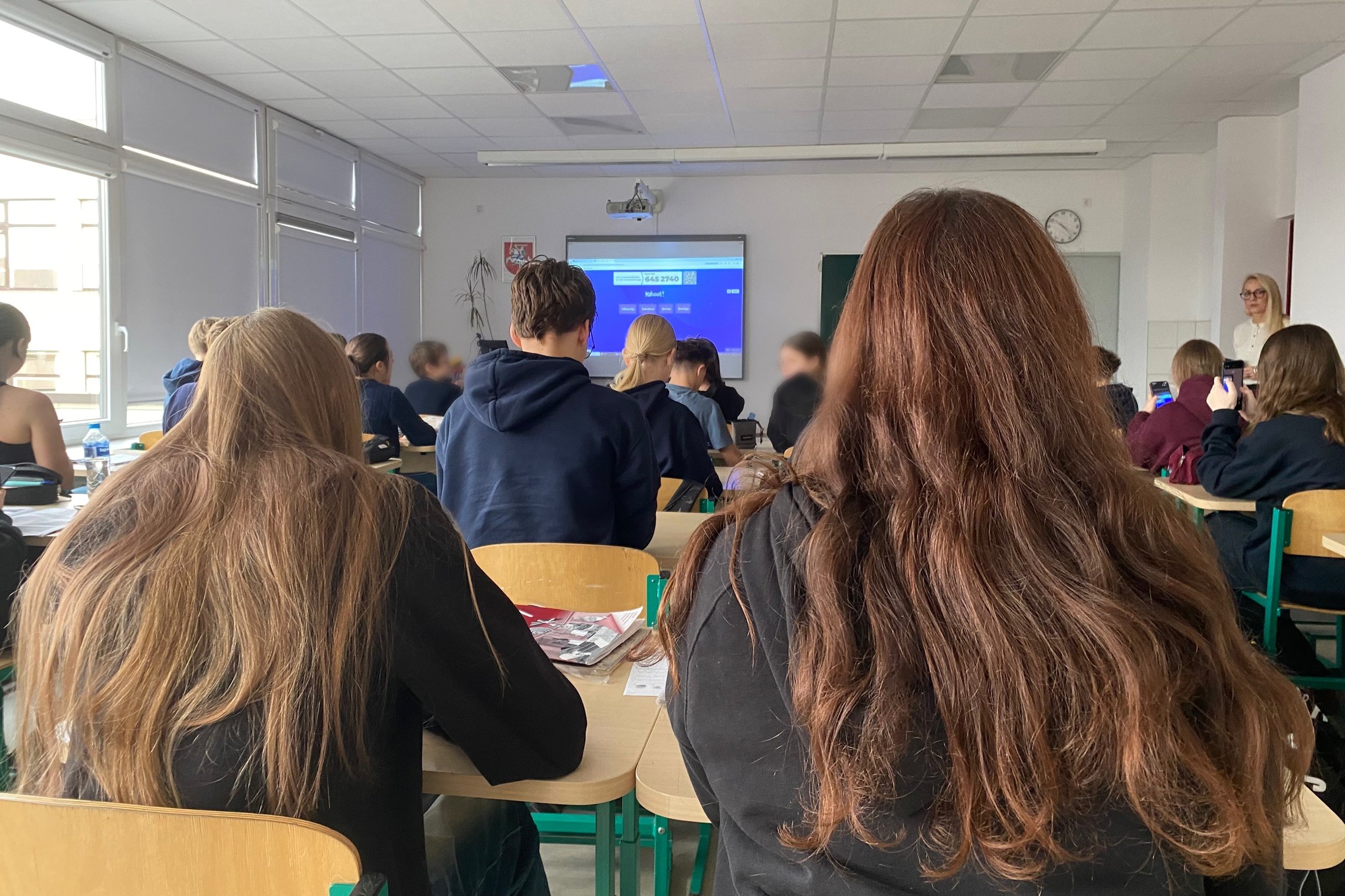 Klasės vaizdas iš suolo galo: mokiniai sėdi prie stalų ir žiūri į interaktyvų ekraną, kuriame rodoma skaidrė. Priekyje stovi mokytoja, keli mokiniai laiko telefonus, klasėje matyti šviesūs langai ir sieninis laikrodis.