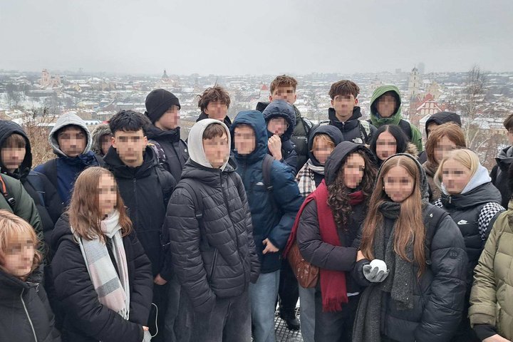 Grupė Kristoforo gimnazijos 9 klasės mokinių, apsirengusių žieminėmis striukėmis, stovi lauke ant apžvalgos aikštelės, už jų matyti sniegu padengtas Vilniaus miesto panoramos vaizdas.