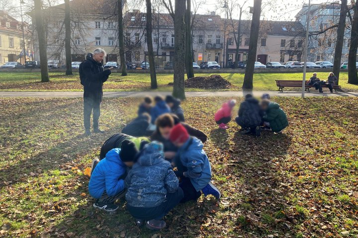 aikų grupė, susiskirsčiusi į kelias mažesnes komandas, atlieka veiklą parke ant nukritusių rudens lapų, o šalia stovi suaugęs žmogus, stebintis ir laikantis rankose užrašų knygelę ar telefoną.
