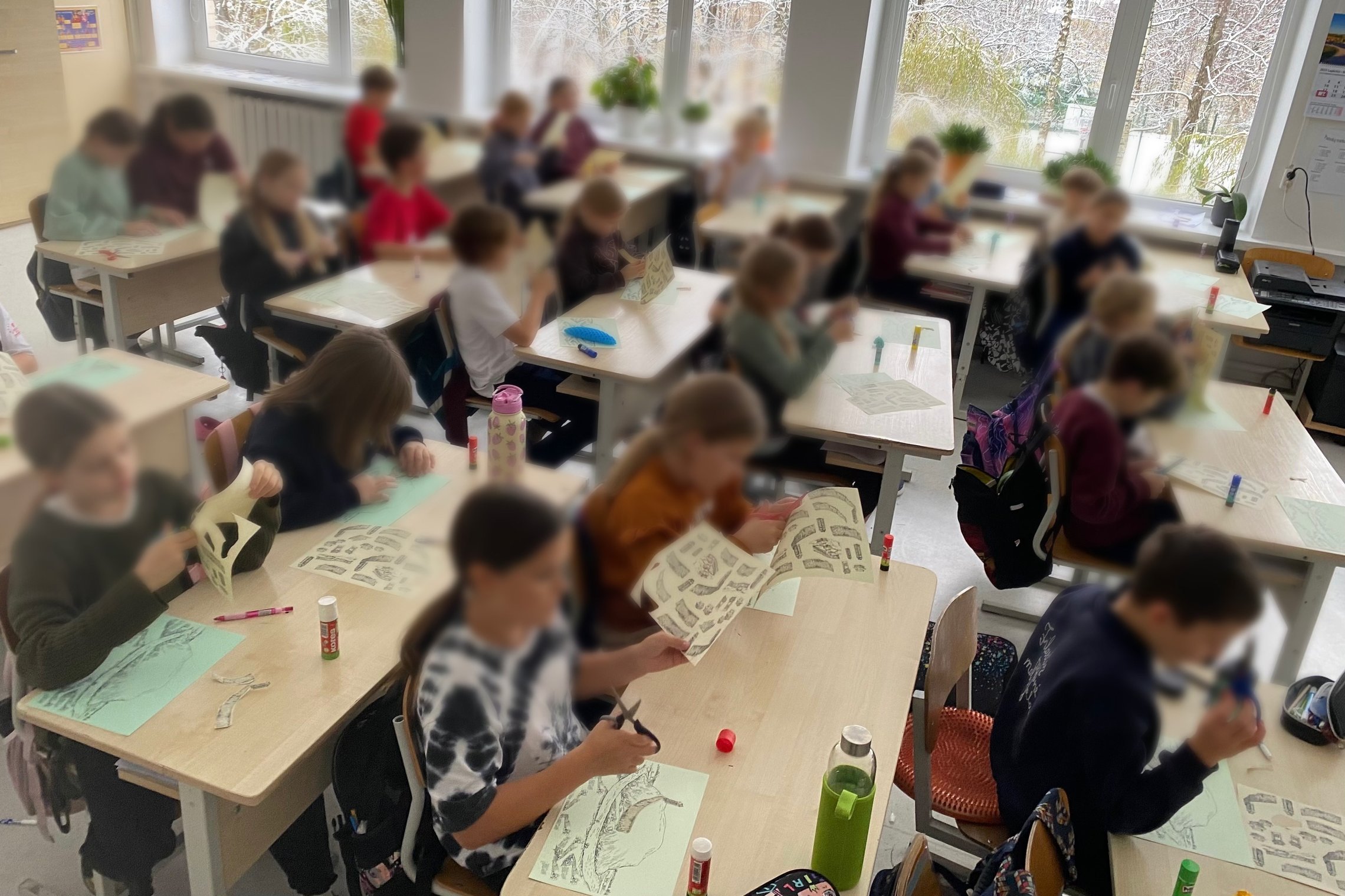 Klasėje prie suolų dirba ketvirtos klasės mokiniai – jie karpo ir klijuoja iliustracijas, kurdami piliakalnių modelius. Vaikai susitelkę į kūrybinę užduotį, ant stalų matyti žirklės, klijai ir popieriaus lapai. Už lango – apsnigtas peizažas.