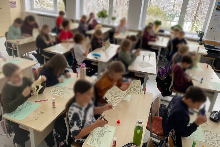 Klasėje prie suolų dirba ketvirtos klasės mokiniai – jie karpo ir klijuoja iliustracijas, kurdami piliakalnių modelius. Vaikai susitelkę į kūrybinę užduotį, ant stalų matyti žirklės, klijai ir popieriaus lapai. Už lango – apsnigtas peizažas.