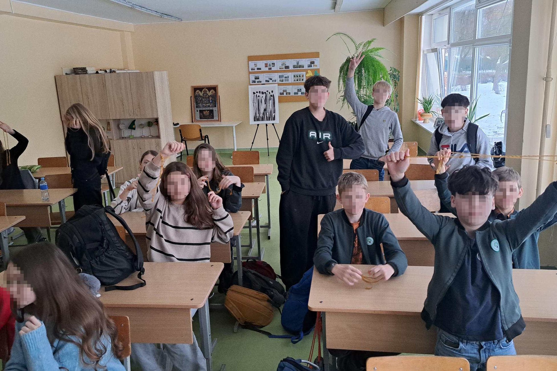 Septintokų grupė klasėje dalyvauja edukacijoje: mokiniai sėdi ir stovi prie suolų, kelia į viršų virveles ar mazgus, o priekyje ir gale matyti kelios žaliuojančios gėlės bei dideli langai, pro kuriuos matosi sniegas lauke.