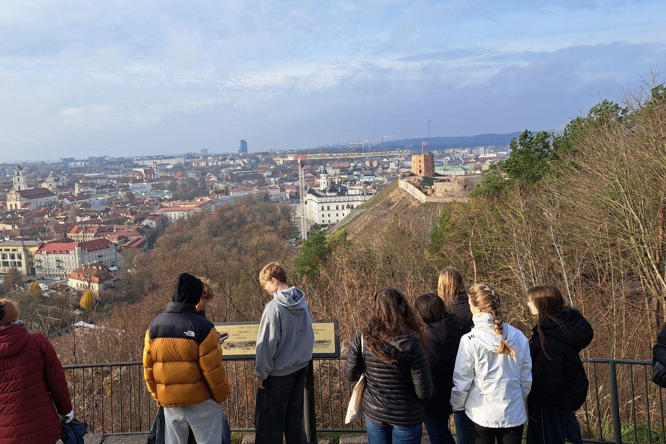Grupė mokinių stovi apžvalgos aikštelėje, žvelgdami į Vilniaus senamiestį; tolumoje matyti Gedimino pilies bokštas ir miesto panorama.