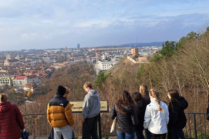 Grupė mokinių stovi apžvalgos aikštelėje, žvelgdami į Vilniaus senamiestį; tolumoje matyti Gedimino pilies bokštas ir miesto panorama.