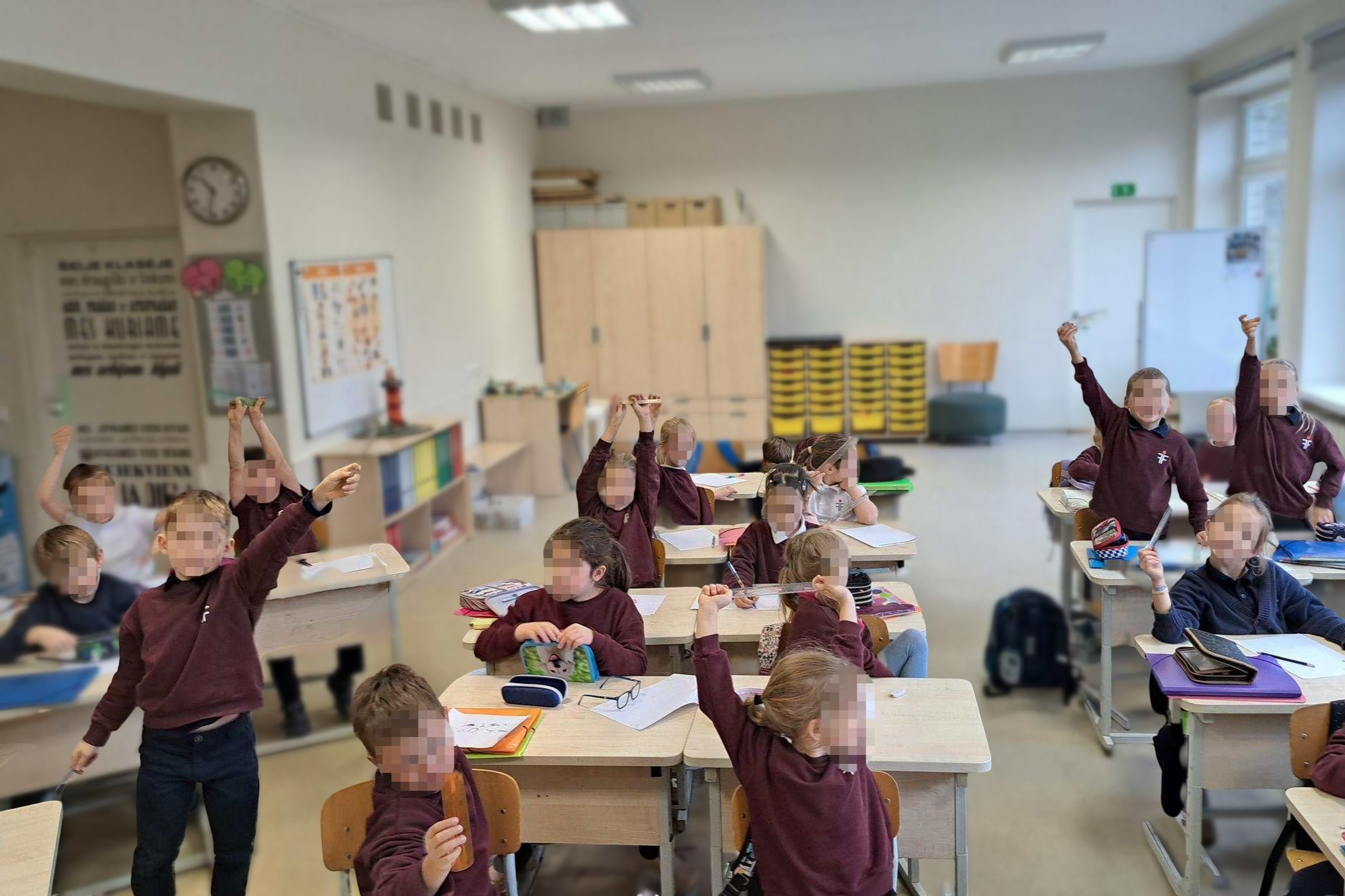 Klasėje pirmokai sėdi prie suolų ir iškėlę liniuotes aktyviai dalyvauja pamokoje; šviesi mokymo aplinka su langais, lentomis ir spintelėmis.