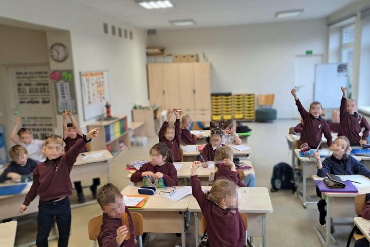 Klasėje pirmokai sėdi prie suolų ir iškėlę liniuotes aktyviai dalyvauja pamokoje; šviesi mokymo aplinka su langais, lentomis ir spintelėmis.