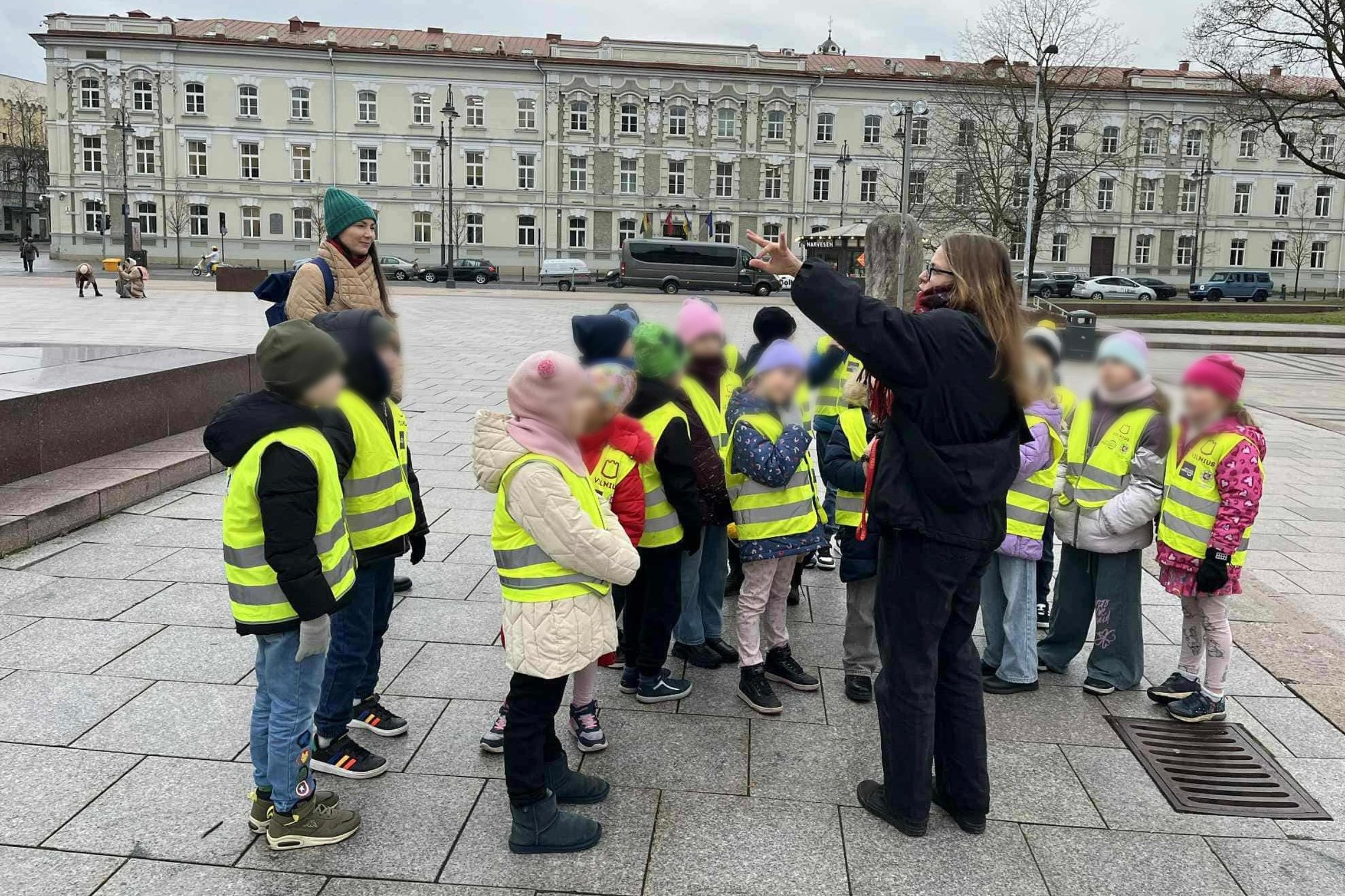 Grupė vaikų su ryškiai geltonomis saugos liemenėmis stovi aikštėje, klausydamiesi suaugusio žmogaus, rodančio ranka į tam tikrą objektą