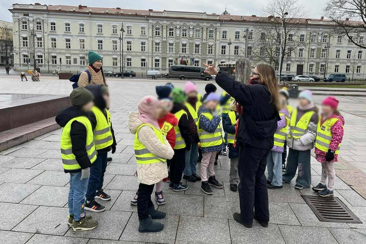 Grupė vaikų su ryškiai geltonomis saugos liemenėmis stovi aikštėje, klausydamiesi suaugusio žmogaus, rodančio ranka į tam tikrą objektą