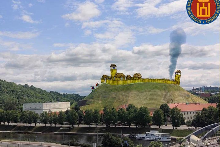 Vilniaus Gedimino kalno nuotrauka, kurioje vizualiai atkurtas senovinis medinis piliakalnio įtvirtinimas su bokštais ir gynybine siena; virš vieno bokšto kyla dūmai, viršutiniame dešiniajame kampe matomas Vilniaus pilių direkcijos logotipas.