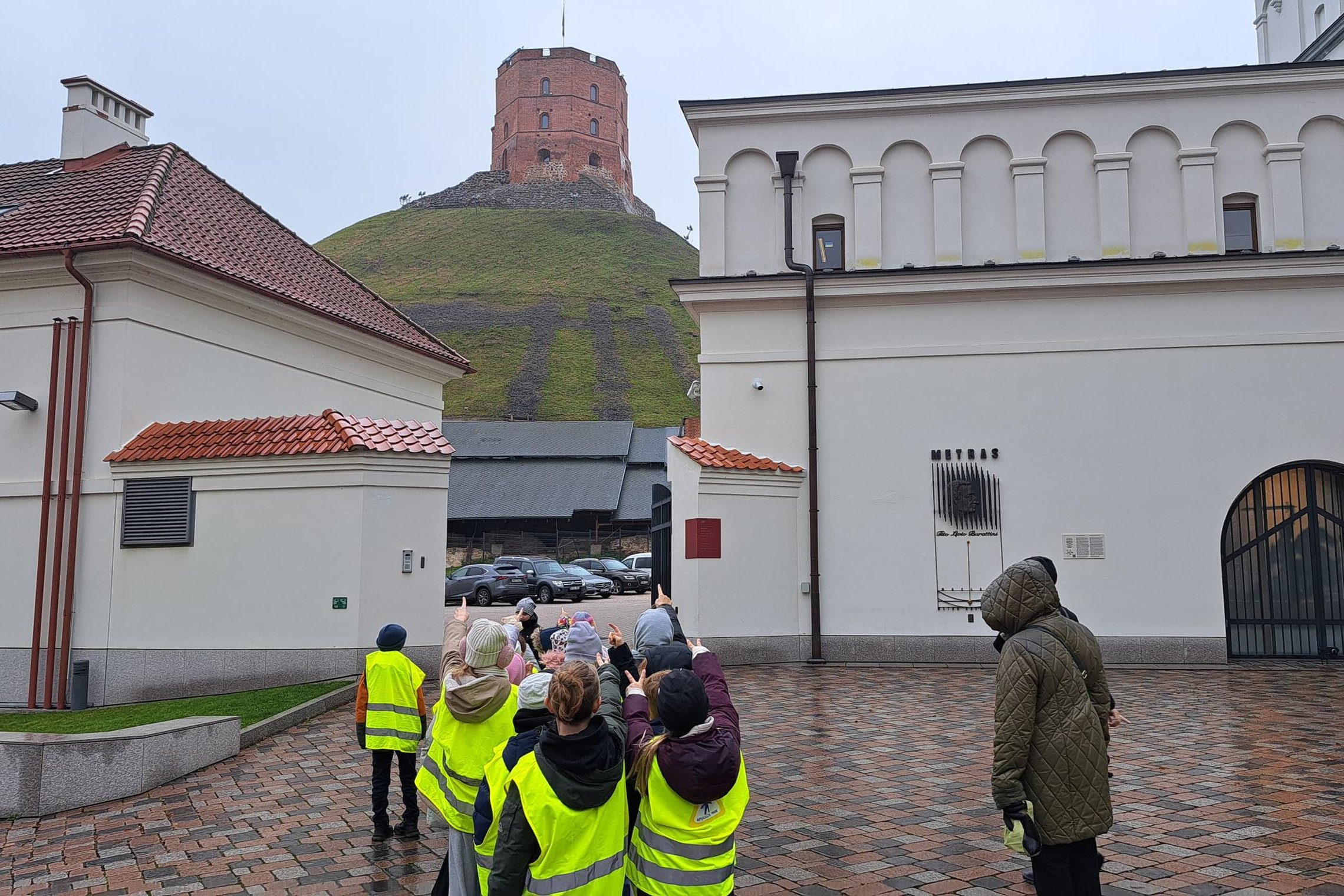 Vaikai su ryškiai geltonomis liemenėmis stovi Katedros aikštėje ir rodo į Gedimino pilies bokštą, matomą ant kalno už baltų pastatų