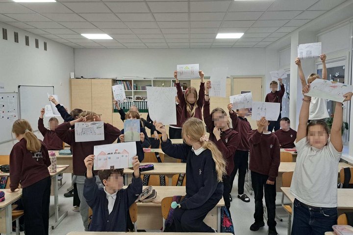 Klasėje stovintys ir sėdintys mokiniai pakeltose rankose laiko savo piešinius, vaizduojančius pilis ir kitus kūrybinius darbus; matyti stalai, kėdės ir klasės įranga.