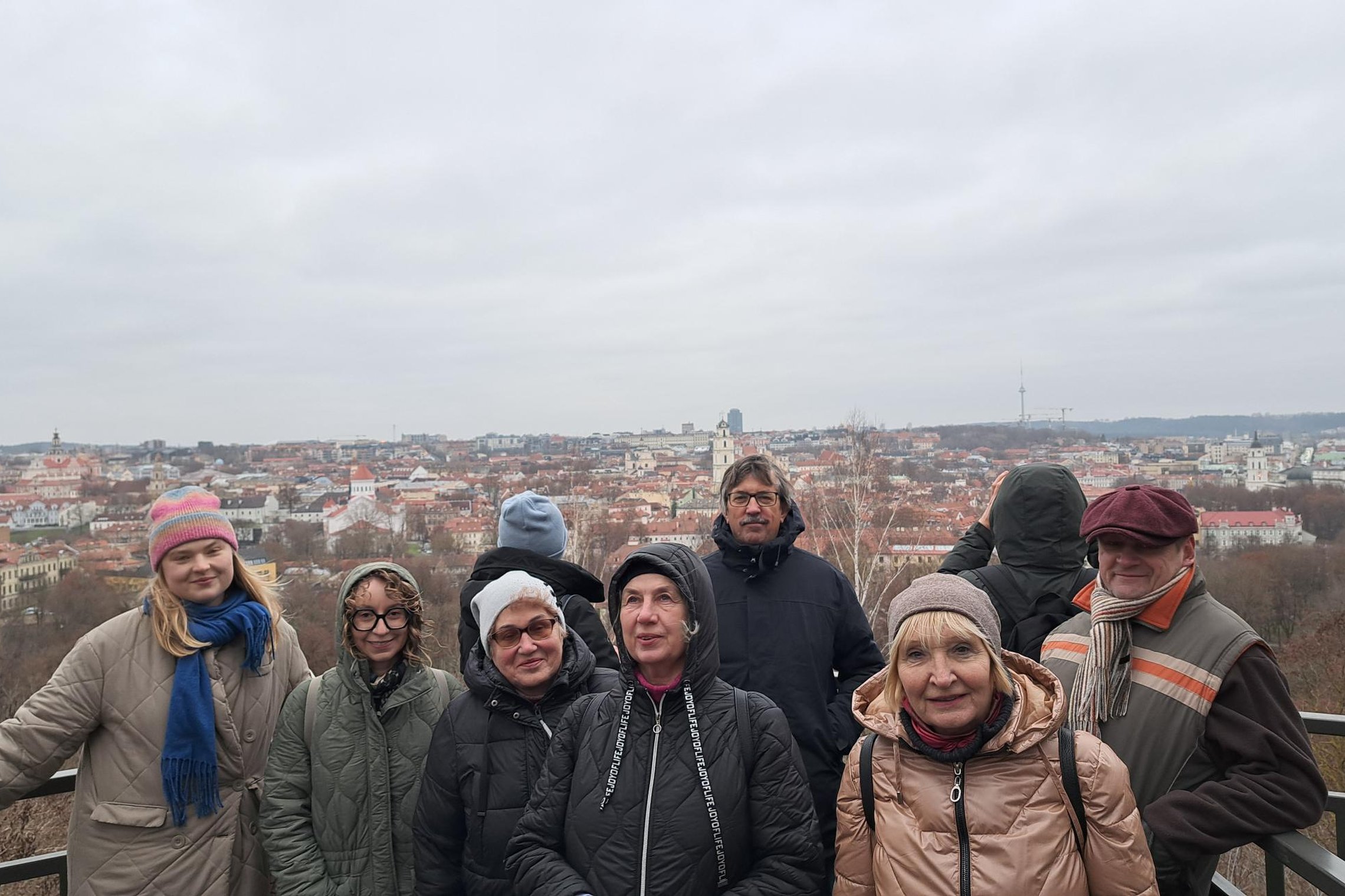 Grupė žmonių, apsirengusių žieminiais drabužiais, stovi Altanos bokštelio apžvalgos aikštelėje; už jų atsiveria plati, apsiniaukusio dangaus apšviesta Vilniaus senamiesčio panorama.