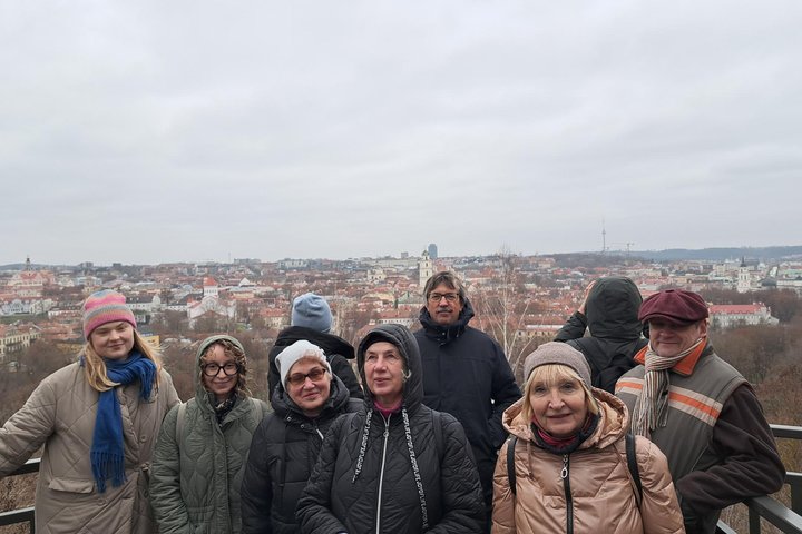 Grupė žmonių, apsirengusių žieminiais drabužiais, stovi Altanos bokštelio apžvalgos aikštelėje; už jų atsiveria plati, apsiniaukusio dangaus apšviesta Vilniaus senamiesčio panorama.