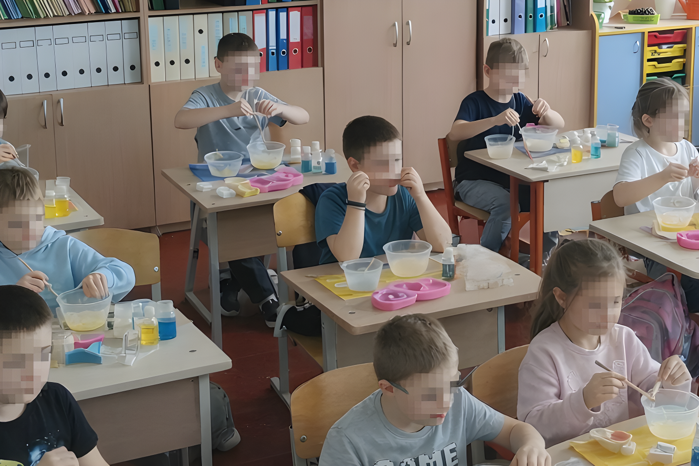 Mokiniai klasėje sėdi prie stalų ir atlieka eksperimentą – maišo skysčius bei medžiagas plastikiniuose indeliuose. Ant stalų matyti mažos buteliukų talpos, pipetės ir spalvoti skysčiai.