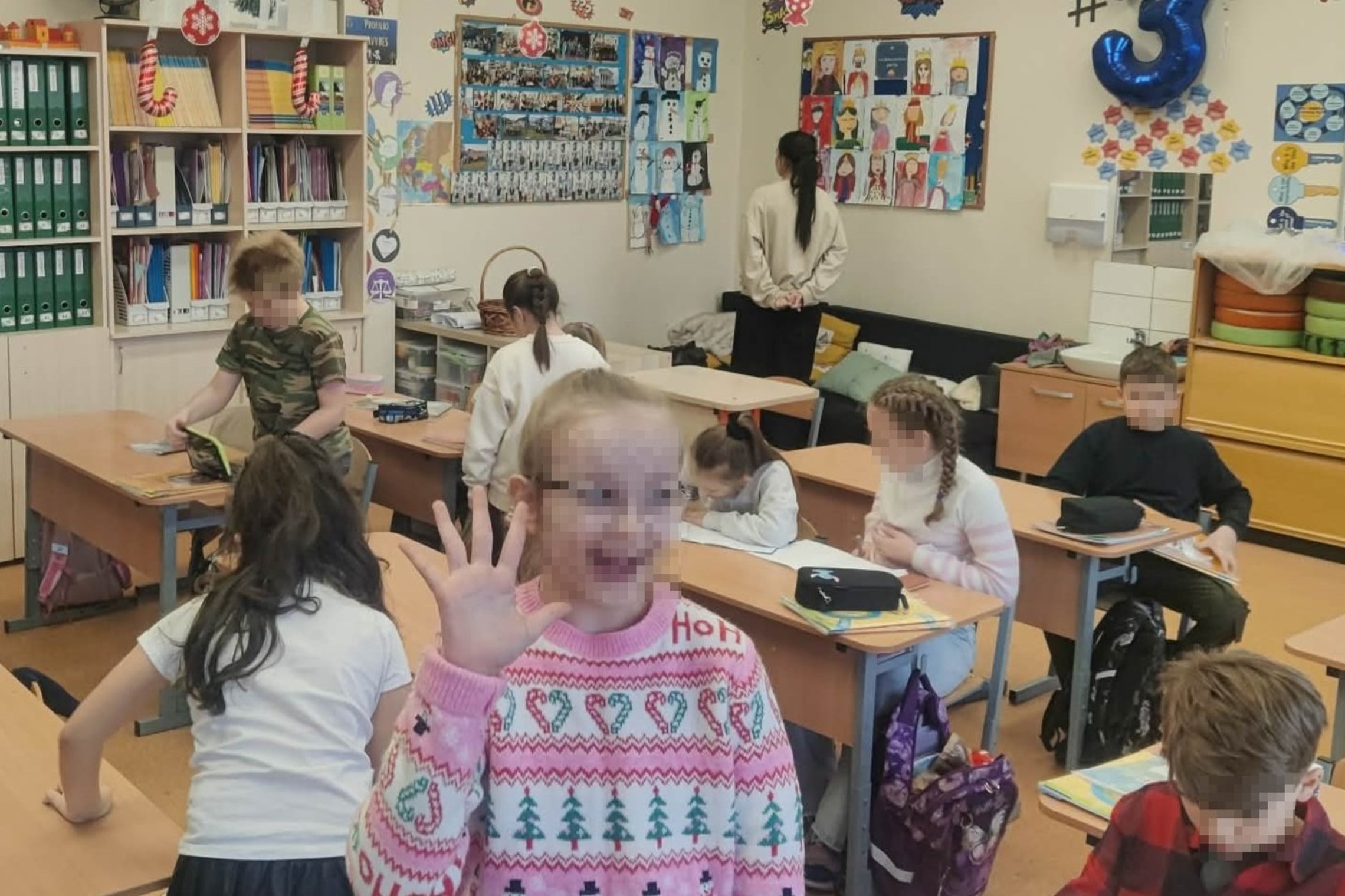 Pradinės mokyklos klasė, kurioje mokiniai sėdi prie suolų ir atlieka užduotis. Priekyje viena mokinė pakėlusi ranką ir šypsosi. Kiti vaikai rašo, piešia arba dirba su pratybomis. Ant sienų matyti vaikų piešiniai, nuotraukų stendai ir mokymosi priemonės, k