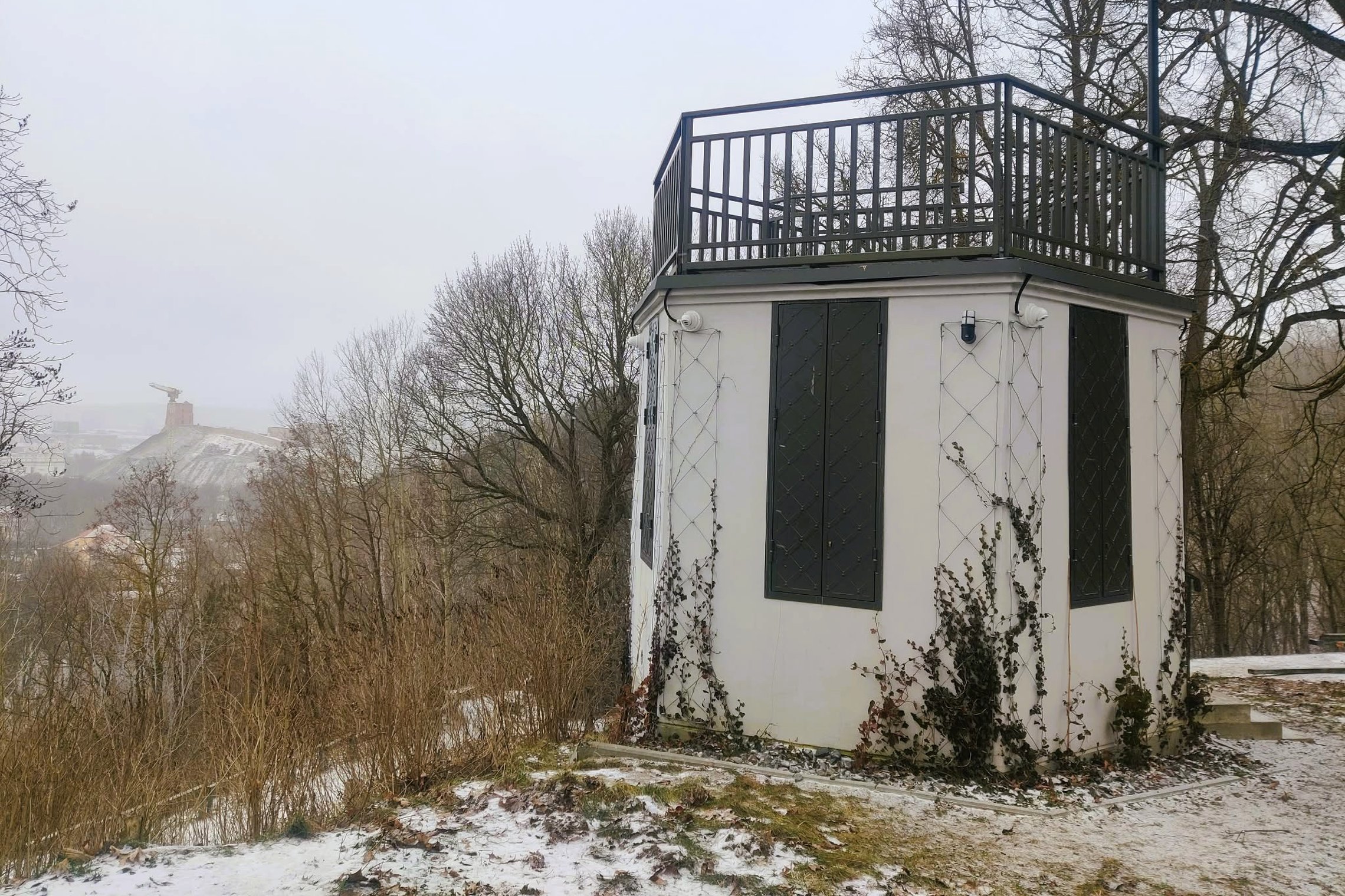 Nedidelis apžvalgos paviljonas ant kalvos su terasa viršuje. Aplink – žiemos peizažas su medžiais ir lengvu sniegu, o tolumoje matyti Gedimino kalnas su bokštu.