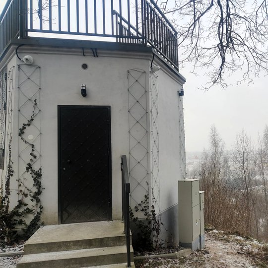 Nedidelis apžvalgos paviljonas ant kalvos su terasa viršuje. Aplink – žiemos peizažas su medžiais ir lengvu sniegu, o tolumoje matyti Gedimino kalnas su bokštu.