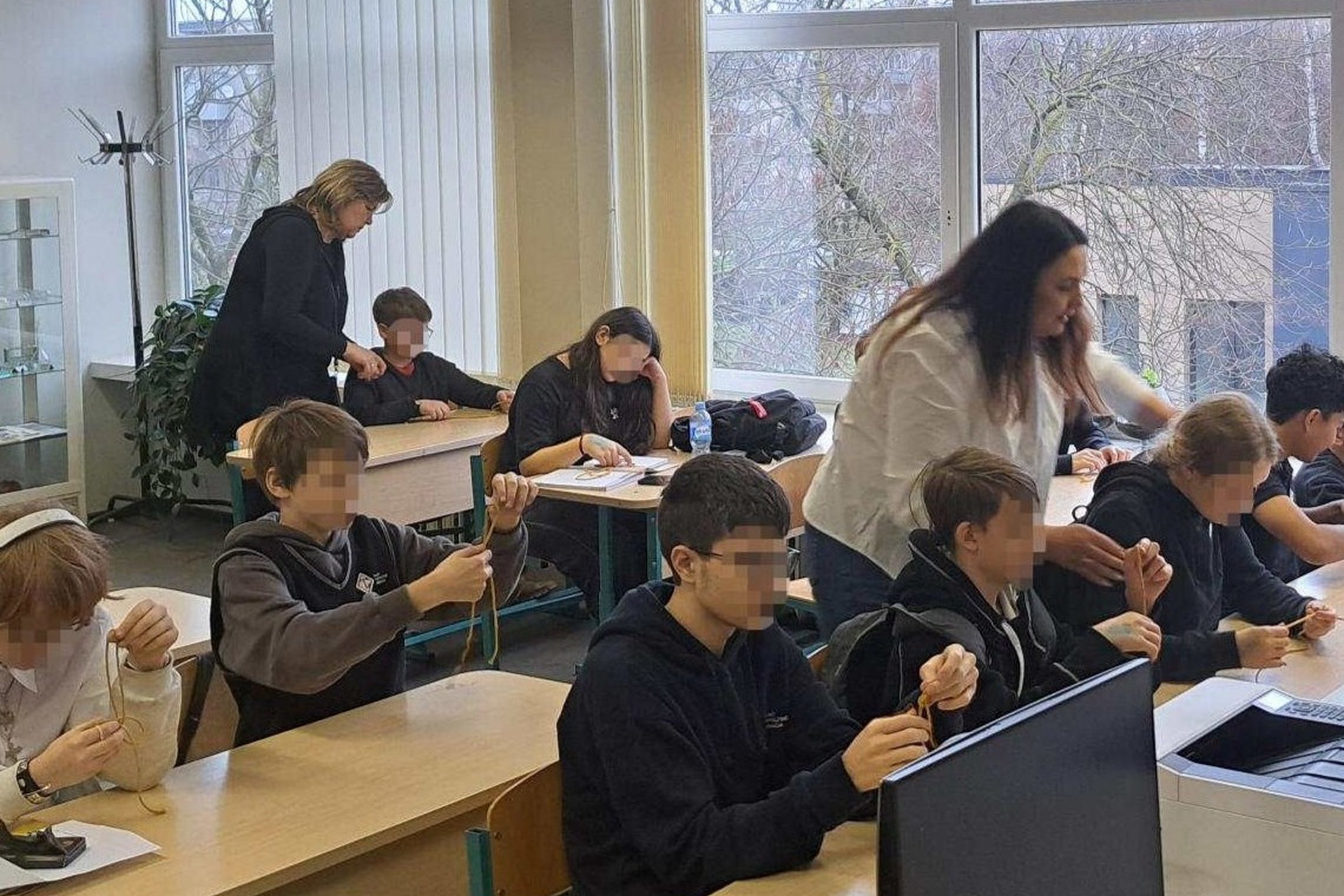 Klasėje prie suolų sėdintys mokiniai pina mazgus, o šalia jų vaikšto ir padeda dvi mokytojos; pro didelius langus matyti žiemos peizažas.