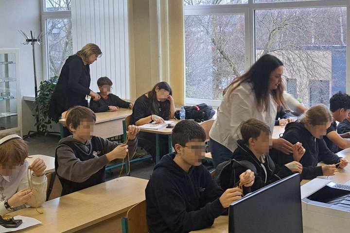 Klasėje prie suolų sėdintys mokiniai pina mazgus, o šalia jų vaikšto ir padeda dvi mokytojos; pro didelius langus matyti žiemos peizažas.