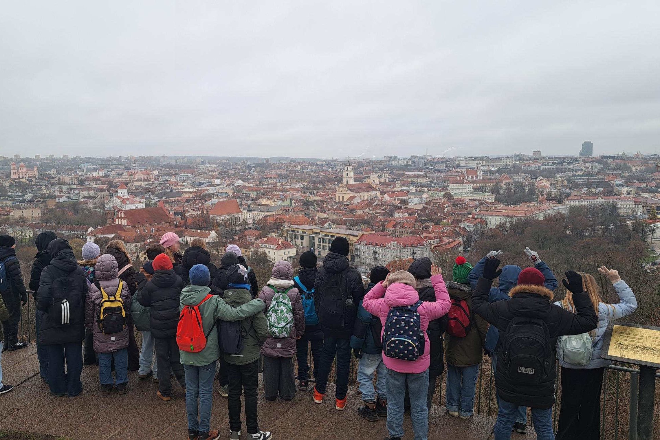 Vaikai žiemos metu stovi apžvalgos aikštelėje ant kalno ir stebi žemiau atsiveriančią Vilniaus senamiesčio panoramą su raudonų stogų pastatais.