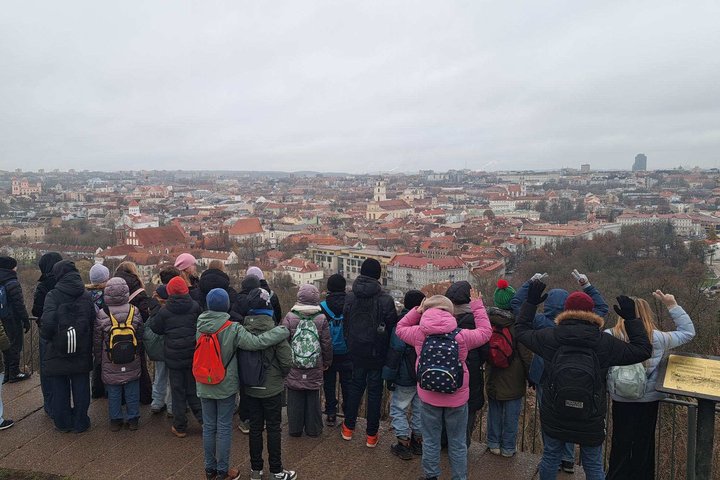Vaikai žiemos metu stovi apžvalgos aikštelėje ant kalno ir stebi žemiau atsiveriančią Vilniaus senamiesčio panoramą su raudonų stogų pastatais.