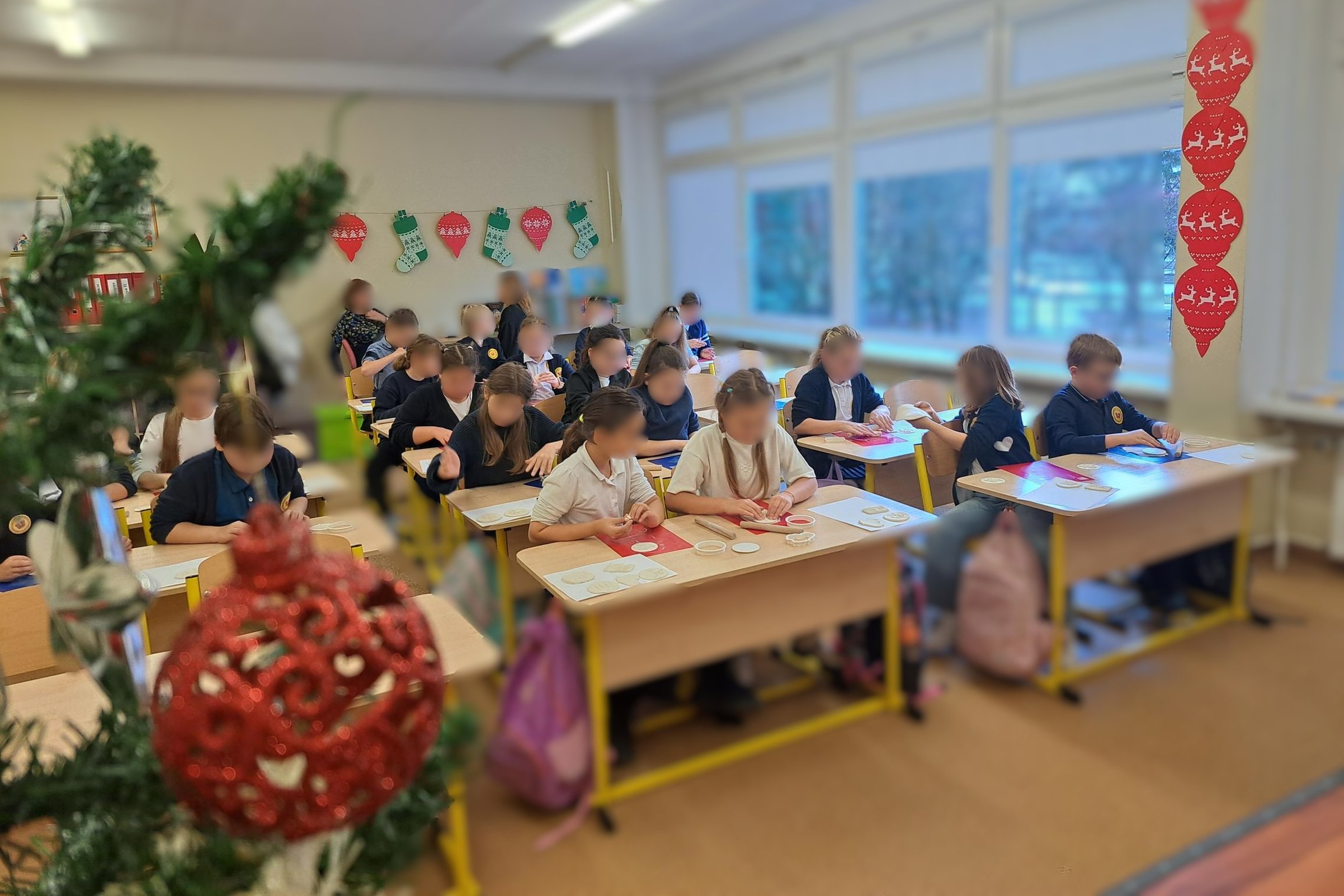 Mokiniai klasėje sėdi prie stalų ir dalyvauja kūrybinėje veikloje – iš molio ar plastilino formuoja apskritas figūras. Ant sienos matyti kalėdinės dekoracijos, o klasę puošia eglės šaka su raudonu žaisliuku.