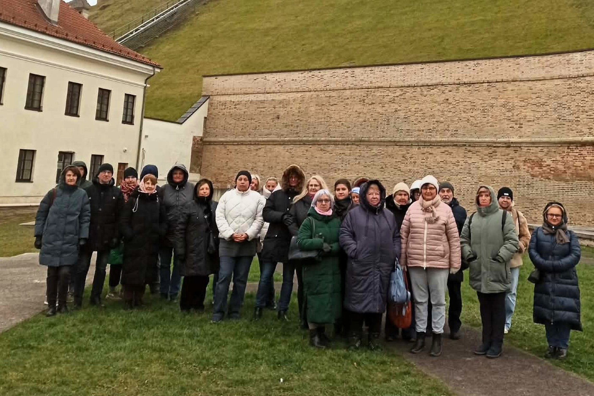 Grupė žmonių stovi Vilniaus pilių teritorijoje prie aukštos plytinės sienos Gedimino kalno papėdėje. Už jų matyti žalias kalno šlaitas ir į viršų kylantis takas.