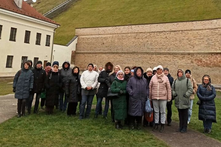 Grupė žmonių stovi Vilniaus pilių teritorijoje prie aukštos plytinės sienos Gedimino kalno papėdėje. Už jų matyti žalias kalno šlaitas ir į viršų kylantis takas.