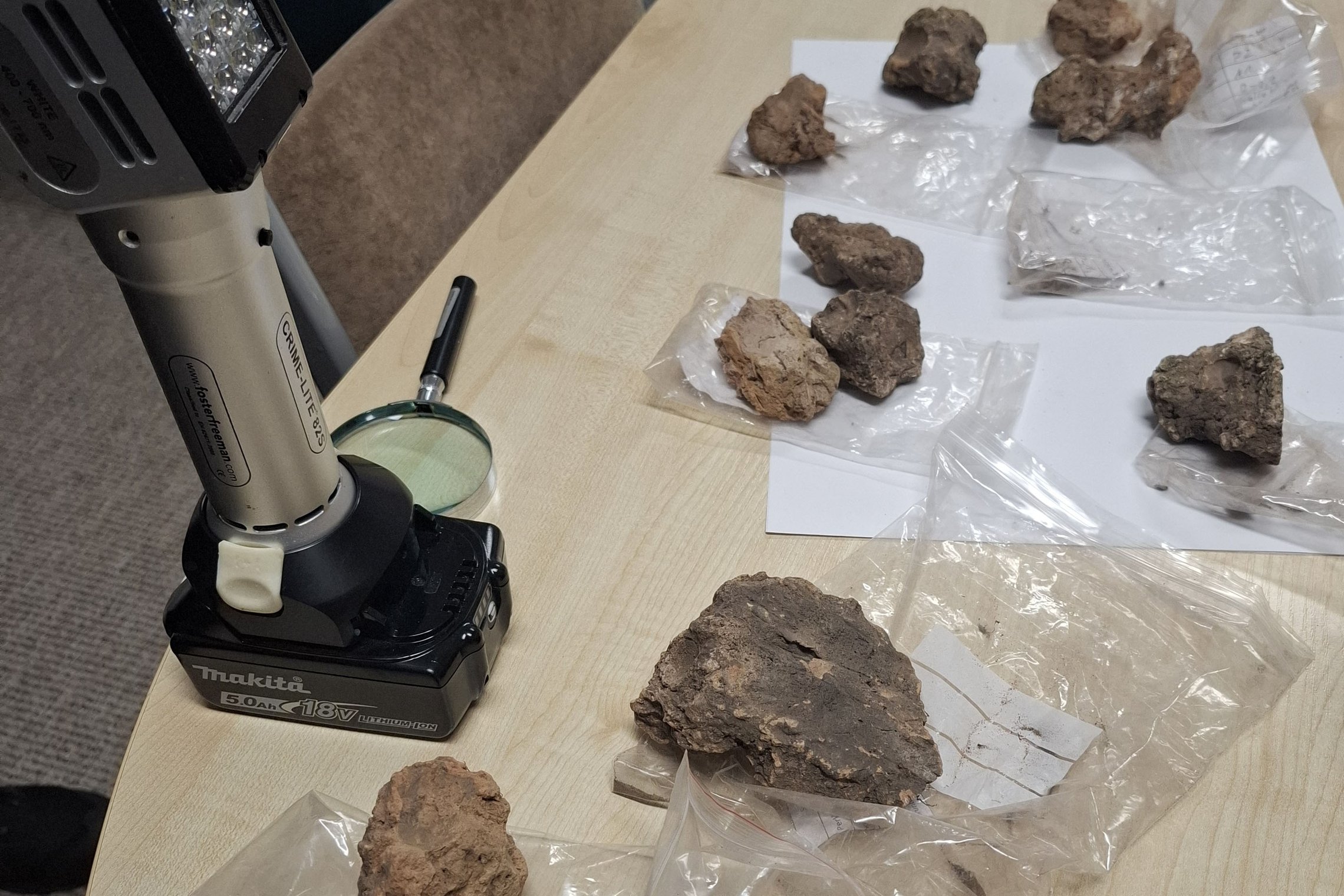 Ant stalo išdėlioti žemės ar molio gabalai plastikiniuose maišeliuose su etiketėmis. Šalia padėtas nešiojamas LED šviestuvas ir didinamasis stiklas, naudojami radiniams apžiūrėti.