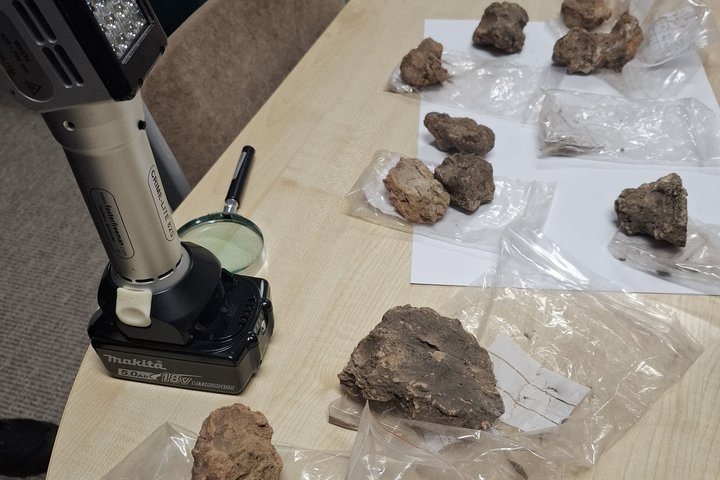 Ant stalo išdėlioti žemės ar molio gabalai plastikiniuose maišeliuose su etiketėmis. Šalia padėtas nešiojamas LED šviestuvas ir didinamasis stiklas, naudojami radiniams apžiūrėti.