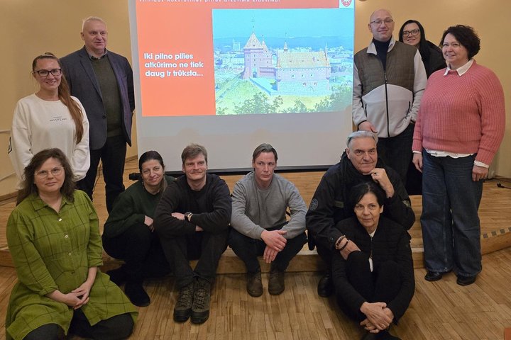 Grupė žmonių pozuoja patalpoje prieš projekcinį ekraną. Ekrane rodoma skaidrė apie Vilniaus Aukštutinės pilies atkūrimą su pilies iliustracija ir tekstu „Iki pilno pilies atkūrimo ne tiek daug ir trūksta…“. Dalis žmonių stovi, kiti sėdi ant grindų priekyj