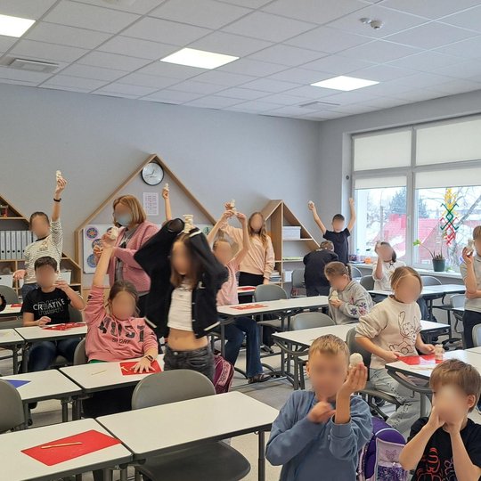 Klasėje vaikai džiaugsmingai kelia į viršų savo iš molio nulipdytus darbelius – figūrėles ar bokštelius. Dalis mokinių stovi, kiti sėdi prie baltų stalų, tarp jų – ir mokytoja. Aplink matyti knygų lentynos, laikrodžiai bei spalvingi vaikų kūriniai ant sie