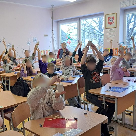 Pradinukai klasėje pakėlę rankas demonstruoja savo nulipdytus molio kūrinius. Vaikai sėdi prie tvarkingai išdėliotų stalų, padengtų spalvotais padėklais ir molio formavimo įrankiais. Fone matyti lentynos su dėžėmis ir segtuvais, spalvingos edukacinės deko