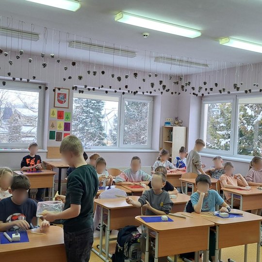 Šviesi pradinių klasių mokinių klasė, kurioje vyksta kūrybinė veikla – vaikai dirba su moliu. Mokiniai sėdi po du ar vieni atskiruose suoluose, ant stalų – mėlyni kilimėliai ir lipdymo įrankiai. Virš galvų nuo lubų kabo kūgiai, pakabinti ant baltų siūlų, 