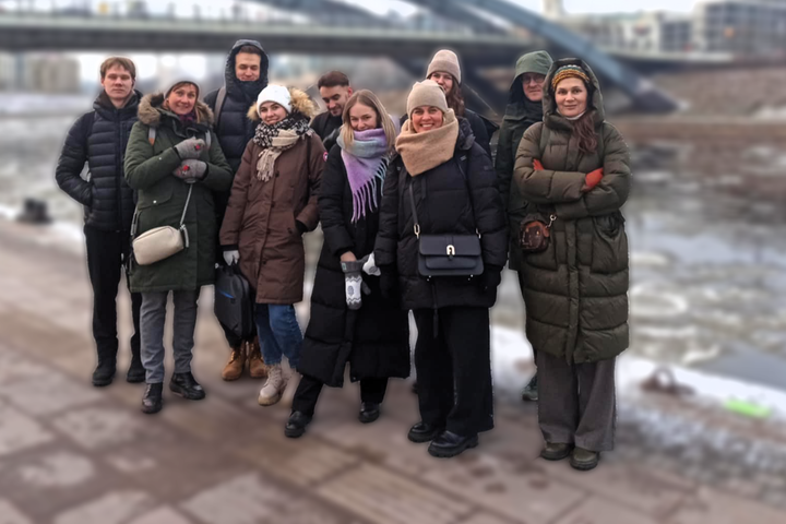 Grupė žmonių pozuoja lauke ant sniegu padengto šaligatvio prie upės. Jie šiltai apsirengę žieminiais drabužiais: paltai, striukės, kepurės ir šalikai. Fone matyti pėsčiųjų tiltas per užšalusią ar ledų pilną upę, matosi pastatai. Visi žmonės atrodo besišyp