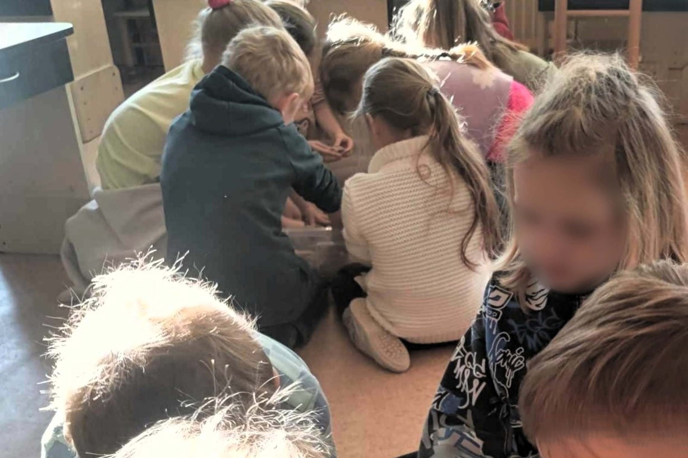 Vaikai susibūrę aplink permatomą dėžę, tyrinėja ir bendradarbiauja su klasės draugais. Jie atrodo įsitraukę į edukacinę veiklą  – stato iš kinetinio smėlio piliakalnius. Ši veikla skatina smalsumą ir bendradarbiavimą.