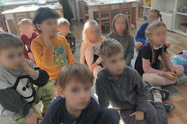 Nuotraukoje matoma grupė vaikų, susėdusių ant grindų šviesioje klasės patalpoje. Jie atrodo susikoncentravę į mokytojos ar vadovo pateiktą užduotį ar pasakojimą. Vaikai įvairaus amžiaus, vilki spalvingus kasdieninius drabužius, o kambarys yra šiltai apšvi