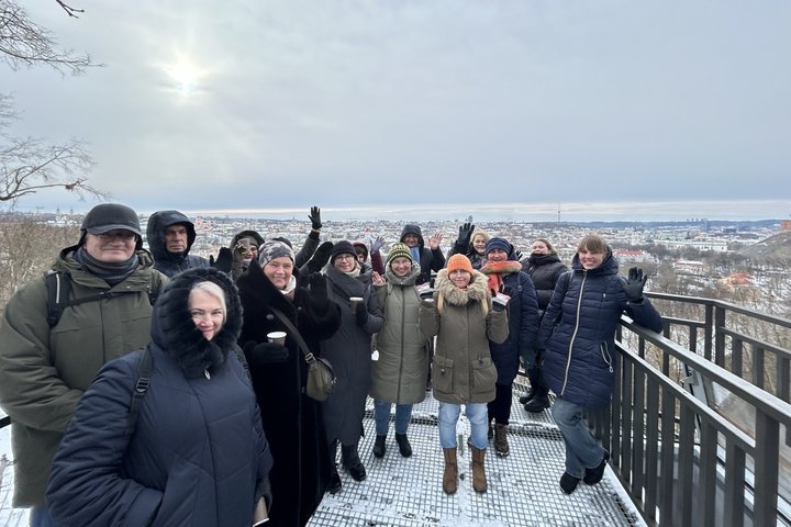 Nuotraukoje matoma grupė suaugusių žmonių, švenčiančių ar dalyvaujančių ekskursijoje, stovi ant apžvalgos aikštelės su žiemos Vilniaus miesto panoramą fone. Visi dėvi šiltus žiemos drabužius ir šypsojasi į kamerą, rodydami pozityvią nuotaiką. Scena fiksuo
