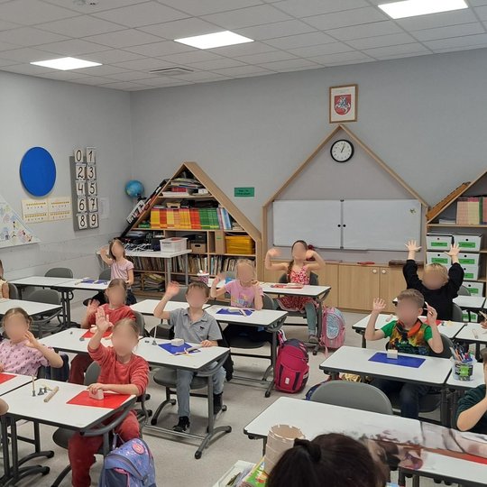 Moderni, šviesi pradinukų klasė, kurioje vaikai kuria darbelius iš molio. Mokiniai sėdi atskiruose baltuose staluose, ant kurių padėti spalvoti kilimėliai, lipdymo įrankiai ir molio gabalėliai. Dalis vaikų pakėlę rankas, kai kurie šypsosi ir mojuoja. Klas