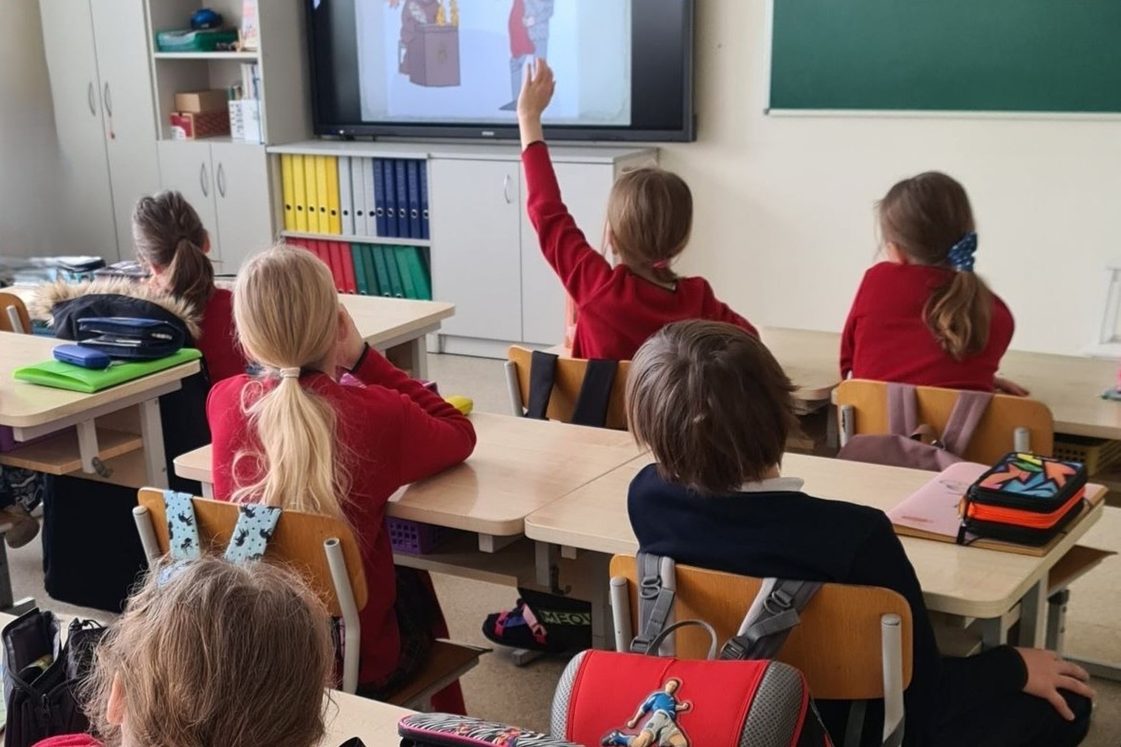 Nuotraukoje penkios mergaitės ir vienas berniukas sėdi klasėje prie stalų. Mergaitės vilki raudonus džemperius, berniukas – tamsiai mėlyną. Viena mergaitė priekyje kelia ranką į viršų, o prieš ją ir kitus vaikus yra televizorius su paveikslėliais. Ant kai