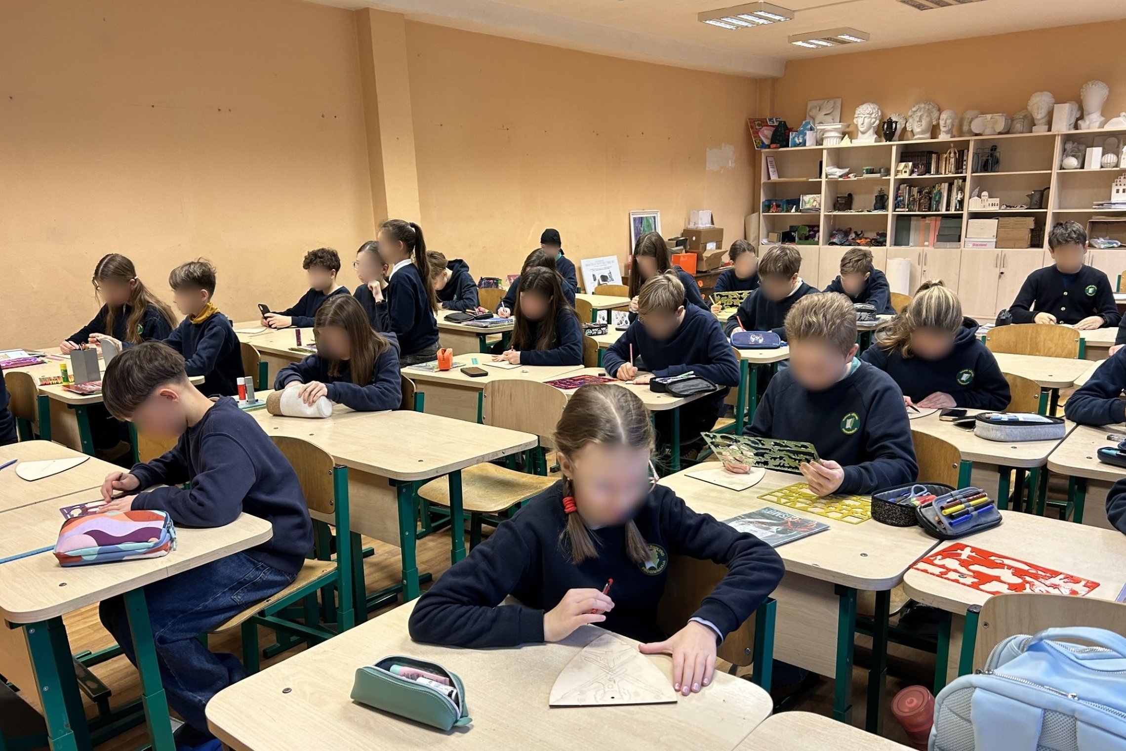 Nuotraukoje pavaizduoti mokiniai, sėdintys klasėje. Visų jų veidai yra sulieti. Visi iš jų yra susifokusavę į kokį nors dalyką, esančio jiems po ranka. Daugumai tai sąsiuvinys ir kažką rašosi, kai kurie laiko liniuotes, o vienas vaikas klasėje sėdi su tel