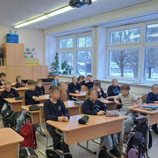 Nuotraukoje matoma klasė, kurioje vaikai sėdi prie suolų ir žiūri į priekį. Jų veidai paslėpti. Klasės gale stovi spintos, ant jų padėti kūrybiniai darbai – dekoracijos su skaičiumi 40 ir kitos spalvingos kompozicijos. Dešinėje pusėje yra dideli langai su
