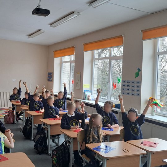 Šviesioje klasėje mokiniai, vilkintys mokyklines uniformas, kelia rankas aukštyn, laikydami įvairius darbelius. Klasės sienos papuoštos mokomaisiais plakatais ir spalvotais popieriniais paukščiais ant langų, pro kuriuos matyti medžiai.