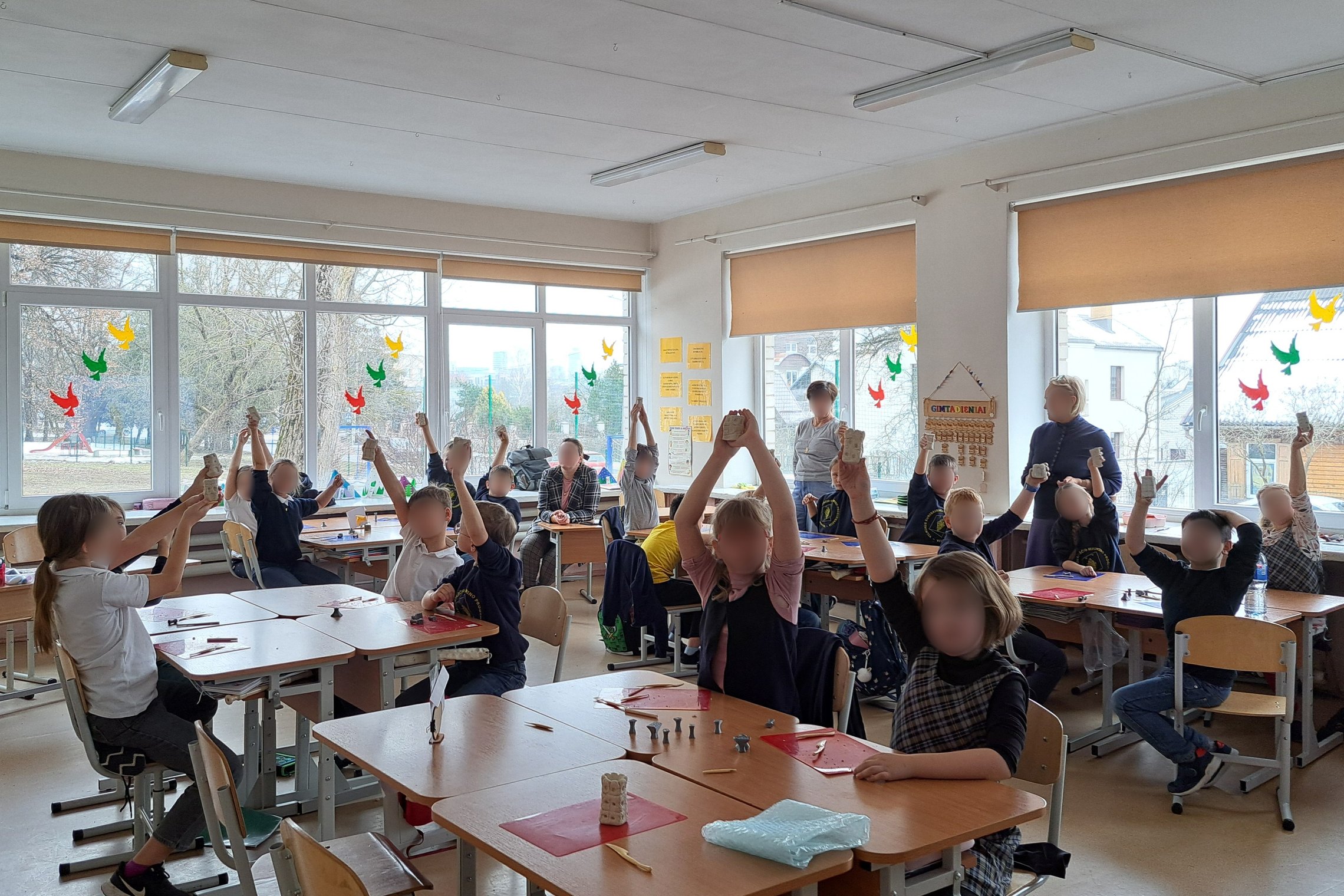Šviesioje klasės patalpoje grupė mokinių kelia rankas aukštyn, laikydami įvairius objektus iš molio. Klasės langai puošti spalvingais popieriniais paukščiais, pro juos matyti parkas ir pastatai. Kambarys pilnas ryškios dienos šviesos ir mokomųjų plakatų a