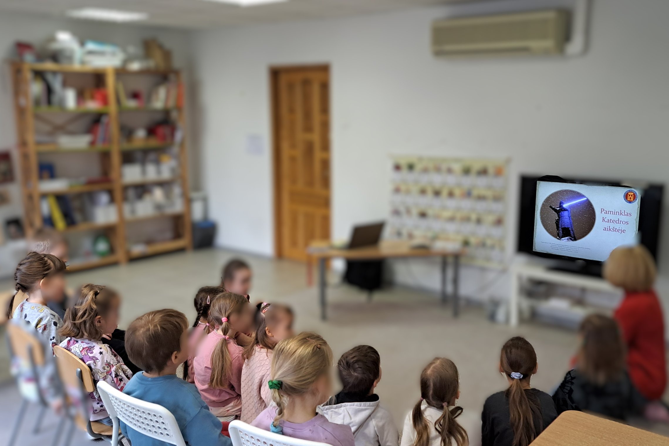Vaikų grupė, sėdinti šviesiame klasės kambaryje, žiūri į televizorių, kuriame rodoma edukacinė medžiaga apie Gedimino paminklą. Vaikai įdėmiai stebi ekraną. Klasėje stovi knygų lentyna su įvairiausiomis priemonėmis.