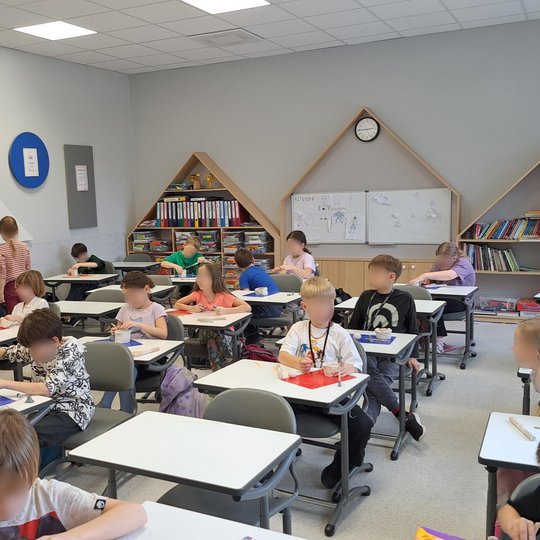 Šiuolaikiška pradinės mokyklos klasė, kurioje vaikai dirba su moliu. Mokiniai sėdi prie baltų individualių stalų, ant kurių išdėlioti spalvoti padėklai, molio gabalėliai ir įrankiai. Keli vaikai atsistoję ar pasilenkę į kūrybinį darbą. Klasėje yra tvarkin