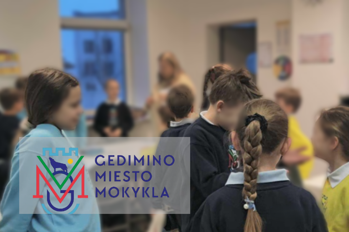 Nuotraukoje matome patalpą, kurioje yra pilną vaikų, amžius yra apie 9-11 metų mokiniai. Nuotraukos priekyje yra išryškinti keturi vaikai, kurie yra nusisukę nuo kameros ir kalbasi tarpusavyje. Fono vaizdas yra susiliejęs, tačiau matosi kiti vaikai. Nuotr