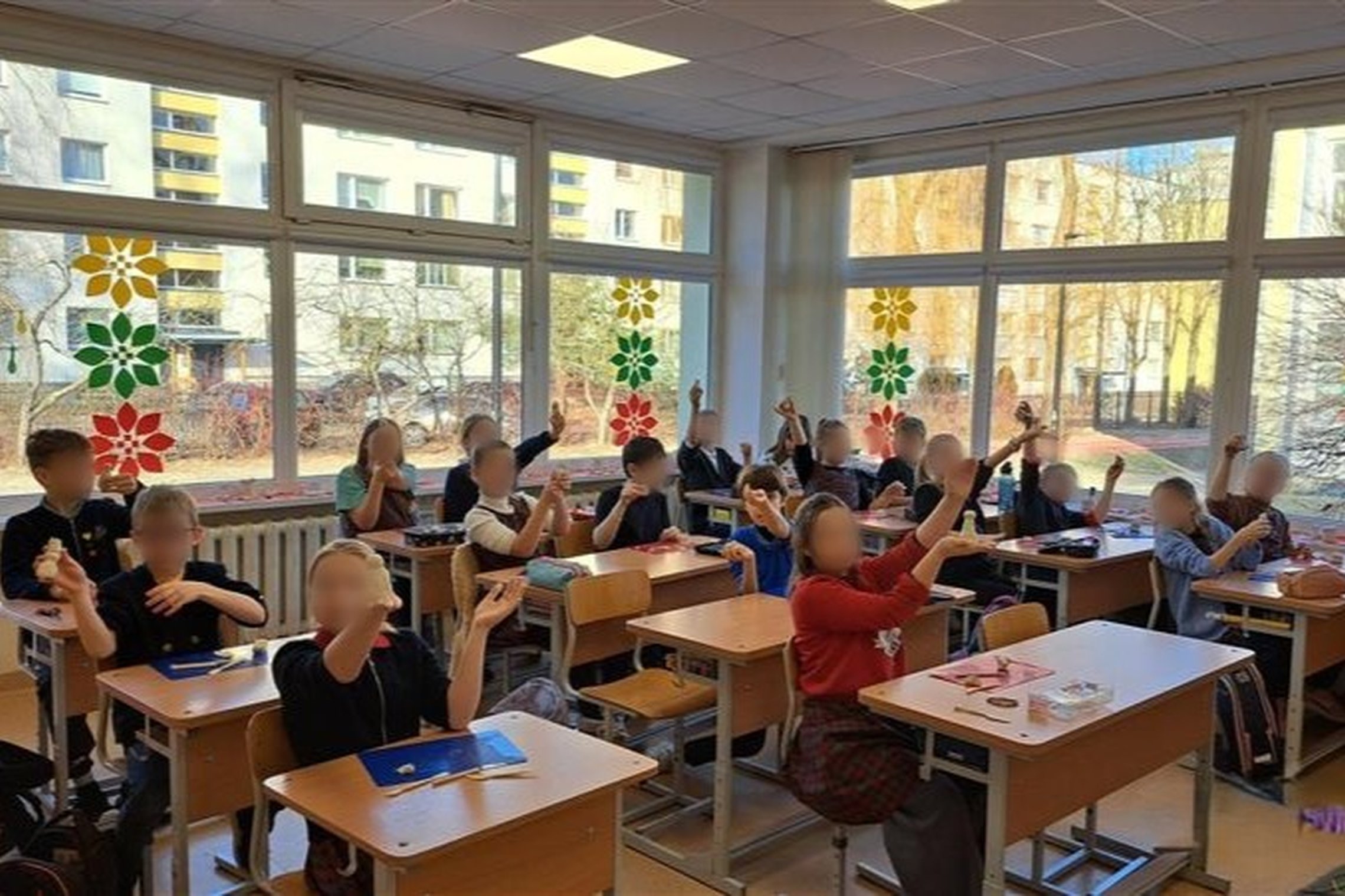 Nuotrauka: klasėje sėdi daug vaikų ir galime suprasti, kad jie kažką gaudo, nes rankos yra ištiestos į viršų. Klasėje yra šviesu, nes už lango šviečia saulė. Ant langų yra užklijuotos Lietuvos trispalvės dekoracijos.