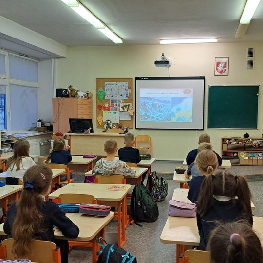 Nuotraukoje vaizduojama klasė, kurioje vaikai sėdi suoluose ir žiūri į priekyje rodomą projekciją su pavadinimu „Ledynams atsitraukiant“. Klasėje šviesu, ant sienų kabo vaikų piešiniai, šalia lentos yra kriauklė ir apvalus veidrodis. Mokinių veidai paslėp
